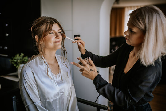 Makeup artist applying blush to smiling bride wearing a silk robe during wedding preparations in a cozy, bright room..
