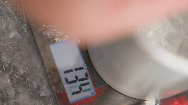 Close up of digital kitchen scale displaying weight reading 142 grams as translucent rubber-like substance is being dropped into metal container placed on scale