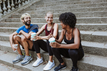 group of multiethnic friends drinking water or coffee and resting after doing exercise together in a City Park in Spain Europe. Hispanic man and afro hair woman wearing athletic clothing