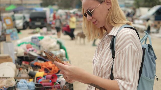 Young blonde traveler wear sunglasses and casual summer outfit browsing wooden souvenirs at bustling outdoor flea market, smiling while exploring local cultural goods and merchandise.Europe travel.