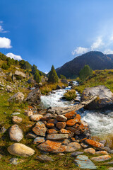 Mountain stream with small wooden bridge, rocky banks, and evergreen trees under a blue sky with clouds.