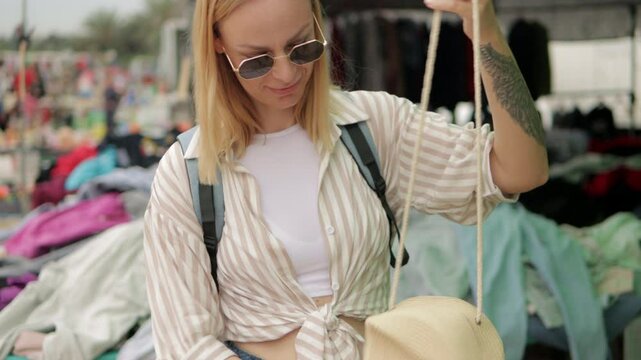 Young woman exploring handmade raffia bag at outdoor flea market, touching woven texture and examining artisan craftsmanship with genuine curiosity while shopping.Europe travel. Lifestyle, vacation,