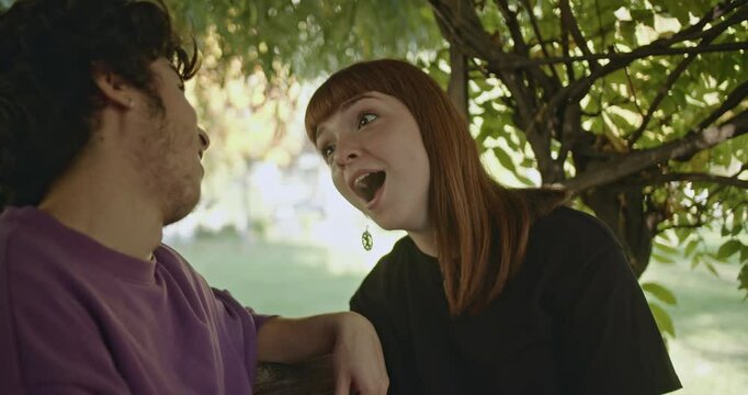 A young couple makes funny faces and playfully teases each other in the park under a tree.