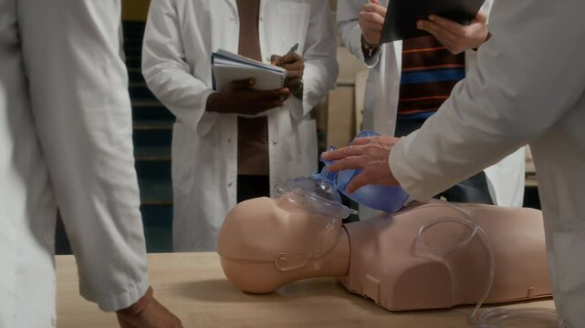 Male professor instructing group of young medical students in cardiopulmonary resuscitation using bag valve mask while demonstrating technique on manikin in training class at university