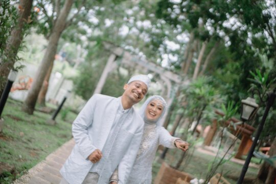 Happy Muslim wedding couple walking and posing in park, wearing traditional white attire. Outdoor Islamic marriage photography, natural and romantic prewedding style.