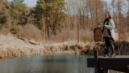 A woman in a beige sweater, black scarf, and polka dot skirt stands on a dock by the water, holding a coffee cup. She gazes at the serene landscape, with a wooden structure and trees in the background