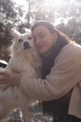 A woman in a beige sweater embraces a white dog, resting her cheek on its head with a peaceful smile. The background is softly blurred, with natural light highlighting the affectionate moment.