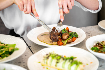 A close-up of hands cutting a piece of meat with a knife and fork on a plate, accompanied by...