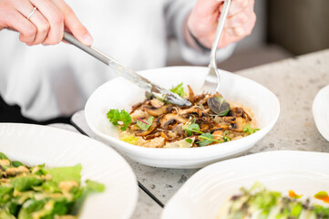 A close-up of hands using tongs to serve a bowl of risotto topped with sautéed mushrooms and herbs. Other dishes are arranged on the table, enhancing the dining experience.