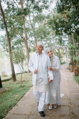 Fototapeta premium Muslim couple wearing elegant white wedding attire, walking together in a lush green garden. Perfect for prewedding, love, romance, and marriage celebration concepts. 