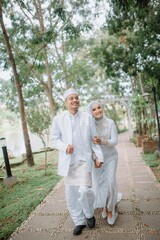 Fototapeta premium Muslim couple wearing elegant white wedding attire, walking together in a lush green garden. Perfect for prewedding, love, romance, and marriage celebration concepts. 
