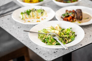 A fresh, vibrant salad served on a white plate, featuring mixed greens, cucumber, and garnished with herbs. Various gourmet dishes are seen in the background on a textured table.
