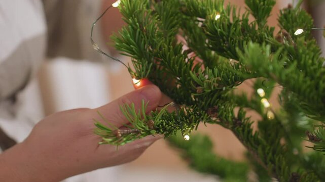 Close up view of hand gently wrapping small glowing string lights around lush green christmas tree branch during cozy holiday decorating session inside warm home interior filled with festive spirit