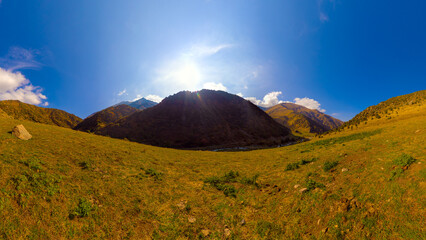 Wide panoramic view of a sunlit mountain valley with grassy slopes under a bright blue sky with...