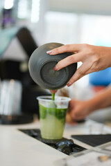 A plastic glass of iced matcha green tea with layer of sparkling water and honey on tea bar counter, selective focus.
