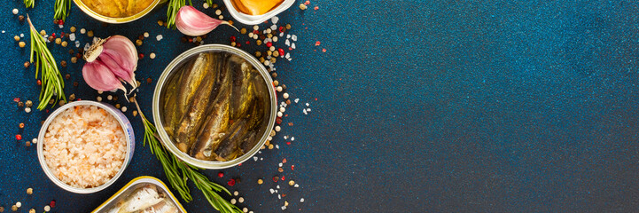 Different open tin cans with canned fish like salmon, mackerel, sprat, sardine, shrimp, among spices and herbs on a blue background, viewed from the top, banner