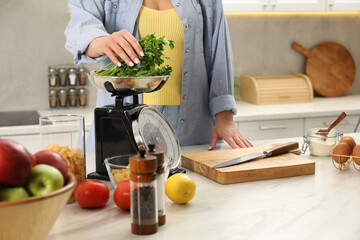 Woman putting parsley into bowl on mechanical kitchen scale at white marble table indoors, closeup