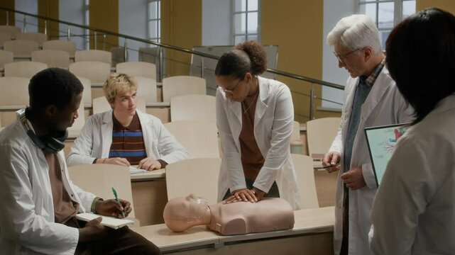 Black teenage girl in white coat practicing chest compression on manikin while male professor making remarks about CPR technique during group training class in auditorium at college