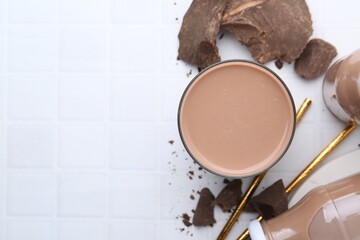 Tasty chocolate milk in glass, bottles and pieces of chocolate on white tiled table, flat lay. Space for text
