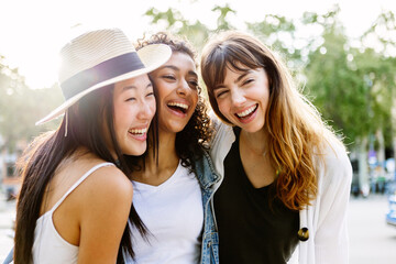Three young smiling women laughing together walking in city street. Diverse female friends having fun together on summer vacation. Youth lifestyle and friendship concept.