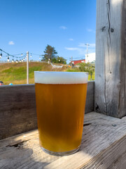 A pint glass filled with a pale ale craft beer. The liquid has a yellow tint. A clear beer glass sits on a table at a microbrewery. The background is the bar area with taps and glasses.