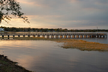 Obraz premium A pier, raised over a calm body of water and juts out from the shore as the evening sun goes down. The wooden structure allows people to fish, moor boats, and dock from the wharf. The sky is golden.