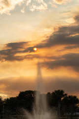A bright golden orange evening sky with clouds and the sun peaking through. The dramatic sunset is over the tops of large trees in a park with a water fountain spraying water into the dramatic sky.
