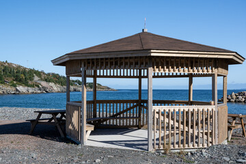 A large octagon-shaped wooden gazebo on a beach overlooking the ocean. The roof is covered with brown shingles. The sides are made of pressure-treated wood. There's a picnic table next to the arbor.