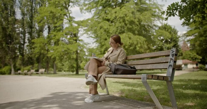 A young businesswoman sits on a park bench and enjoys working remotely using a tablet and notepad in her lap, showcasing the blend of work and leisure