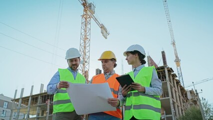 Three engineers in helmets and vests reviewing blueprints and tablet at construction site. Cranes and building framework in background of outdoor project collaboration. Males and female constructors.
