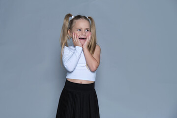 Fototapeta premium little girl with blonde pigtails stands with her hands on her cheeks, showing off a playful, surprised expression. This studio shot captures the whimsy and light-heartedness of childhood, paired with