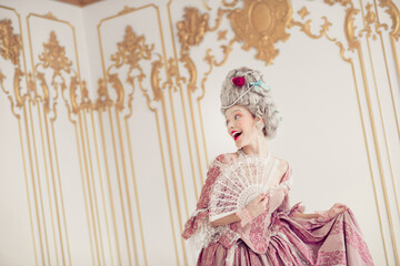 Elegant woman in period attire joyfully holds a fan inside a lavishly decorated historic room,...