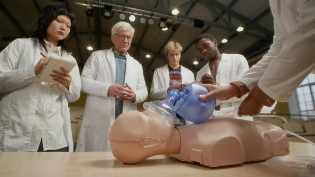 Medical student pumping bag valve mask on manikin practicing cardiopulmonary resuscitation in event of emergency while male professor with study group supervising in training class at college