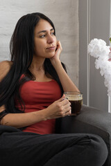 A lovely woman sits on the sofa, sipping coffee, lost in thought while gazing into the distance, in...