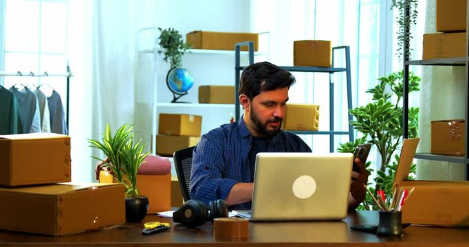 Indian Asian small businessman sitting inside warehouse store surrounded by cardboard boxes on storage rack, typing business report on laptop while checking smartphone for online orders documentation
