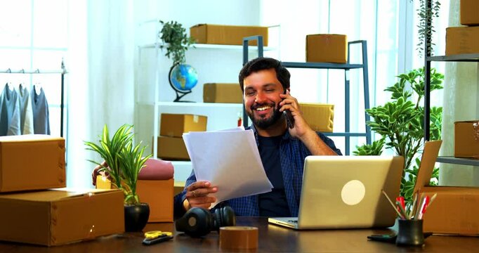 Indian Asian small businessman sitting indoors in storage or warehouse space with cardboard boxes on rack, using laptop and happily talking on phone while holding documents confirming online orders