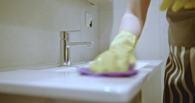 A person wearing yellow gloves and an apron cleans a white bathroom sink with cleaning spray and a purple cloth.