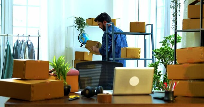 Indian Asian small businessman working in warehouse store room office, picking up cardboard shipment carton box from storage rack and entering product order details on laptop computer at desk