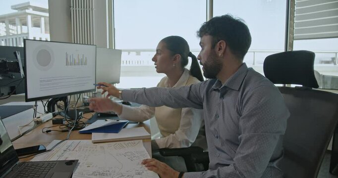 Business colleagues analyze data on computer screens, discussing and pointing at charts, in a modern office setting. - Powered by Adobe