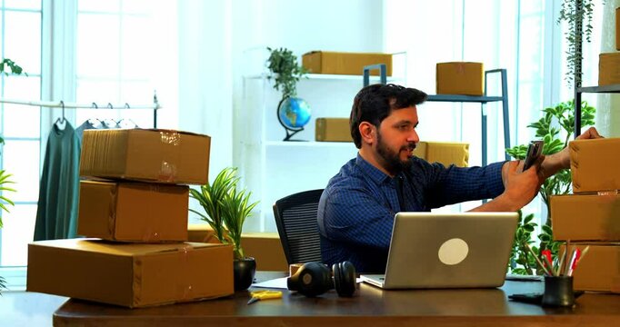 Indian Asian small businessman scanning barcode label on cardboard carton box using smartphone camera while sitting at desk in store room office, working on online order or shipment processing task