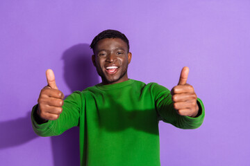 Smiling young man in a green sweatshirt giving thumbs up against a vibrant purple background,...