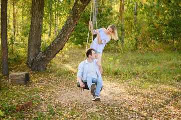 Loving couple swings on tire swing in picturesque place. Man sits on swing, woman stands behind him, swinging together. Loving couple spends time together. Loving couple walks, date.
