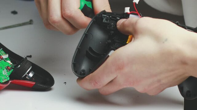 A man in a white shirt repairs a black gaming controller with a small screwdriver. The close-up shot captures his hands and the device against a clean white background.	