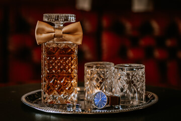 Elegant groom’s accessories with whiskey decanter, bow tie, watch, and crystal glasses arranged on a silver tray in a stylish setting..