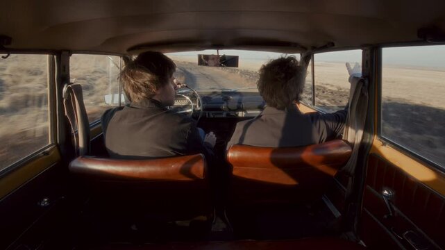Couple sitting in vintage car, driving on country road surrounded by open field, having emotional argument during road trip. Rear view