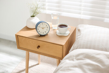 Bedside table with alarm clock, flowers in vase, coffee and glass of water near bed indoors