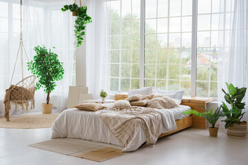 Comfortable bed with lots of soft decorative beige pillows in light sunny bedroom with green plants and large window. Interior design of living room in boho style