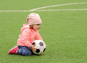 Fototapete Rund Football lesson in kindergarten (preschool) outdoors: little toddler child in pink jacket and hat sitting on green field holding soccer ball. Training and sports for children concept. Copyspace © AMR Studio