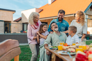 Happy family enjoying backyard barbecue, three generations gathered around table, sharing laughter and food