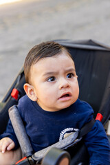Baby in a stroller, sitting with the seat belt on, turning to look at a person while making an angry face